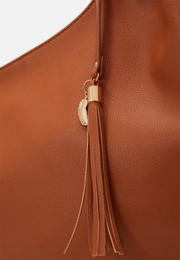 Borsa a tracolla in finta pelle marrone con una texture liscia, un accento in oro con un pompon e un ciondolo a forma di foglia attaccato alla tracolla.