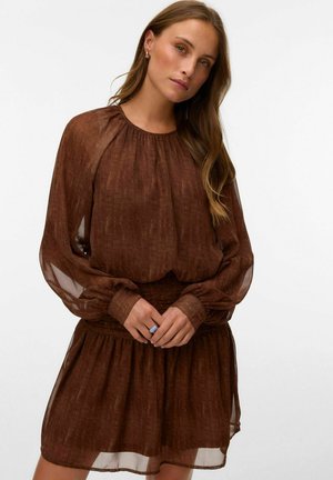 Femme aux longs cheveux bruns portant une robe marron foncé transparente à manches longues avec taille et jupe froncées, debout devant un fond uni.