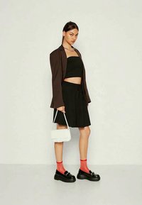 Καφετί σακάκι πάνω από μαύρο crop top και μαύρη φούστα, συνδυασμένο με μαύρα πλατφόρμα παπούτσια και κόκκινες κάλτσες. Κρατώντας μια μικρή λευκή τσάντα.