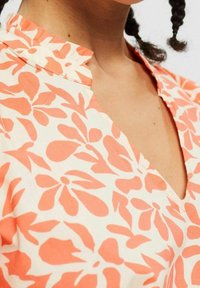 Kurzärmeliges Kleid in Creme mit einem korallenfarbenen Blumenmuster. Mit V-Ausschnitt und Rüschensaum. Hergestellt aus leichtem Stoff.