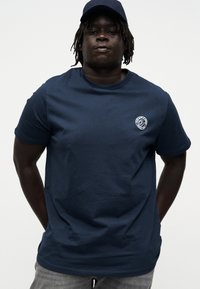 Άνδρας με σκούρο δέρμα και dreadlocks που φοράει σκούρο μπλε καπέλο και σκούρο μπλε T-shirt με μικρό λευκό στρογγυλό λογότυπο στο στήθος.