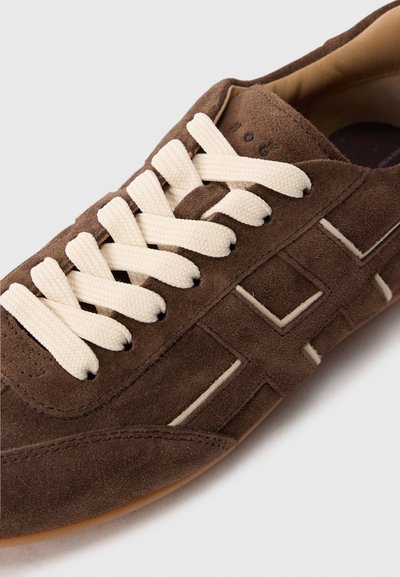 Sneaker in suede marrone con lacci spessi crema, motivi di cucitura geometrici e una fodera interna marrone chiaro.