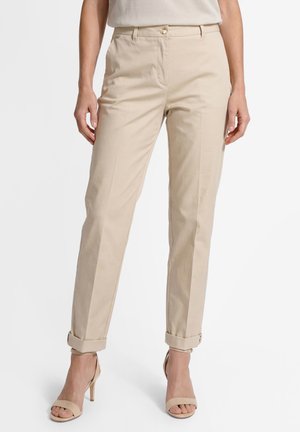 Kobieta ubrana w beżowe, dopasowane spodnie do kostek z podwiniętymi nogawkami oraz beżowe sandały na obcasie z odkrytymi palcami, stojąca na białym tle.