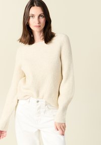 Maglione beige a coste con maniche lunghe, scollo rotondo e texture morbida, abbinato a jeans bianchi a gamba dritta.