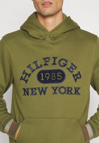 Sudadera con capucha de color verde oliva con el texto bordado "HILFIGER 1985 NEW YORK" en azul marino y amarillo, con puños acanalados a rayas y bolsillo frontal.