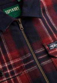 Camisa de franela a cuadros en rojo y azul marino con un cierre dorado, bolsillos laterales y una etiqueta verde. Presenta una tela texturizada y líneas blancas en contraste.