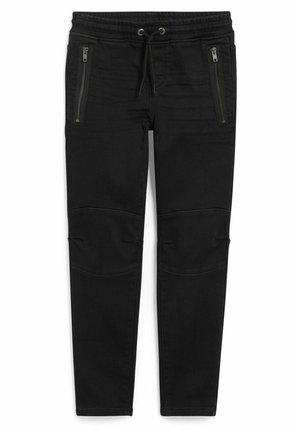 Barn iført en langærmet farveblokskjorte med teksten "New York USA", sorte bukser og hvide sneakers, stående mod en ensfarvet baggrund.