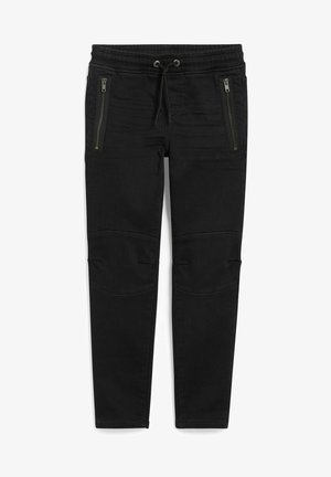 Barn iført en langærmet farveblokskjorte med teksten "New York USA", sorte bukser og hvide sneakers, stående mod en ensfarvet baggrund.