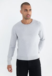Ljust grått texturerat tröja med rund halsringning och ribbade muddar, kombinerad med svarta byxor. Materialet verkar mjukt och stretcht.