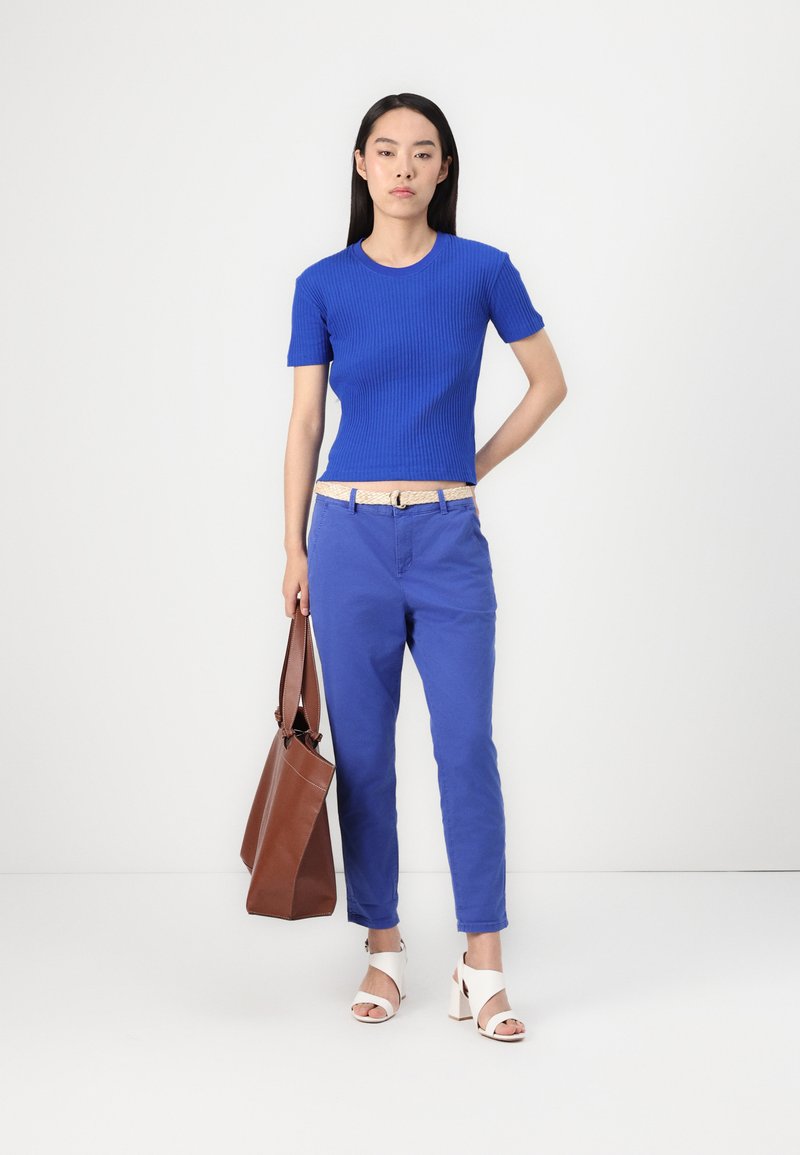 Niebieski prążkowany top z krótkim rękawem połączony z niebieskimi chinosami, beżowym paskiem i brązową torbą. Białe sandały na obcasie dopełniają stylizację.