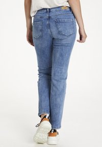 Zili džinsi ar sašaurinātu kāju dizainu, vidēja augstuma jostasvieta, aizmugurējām kabatām un gaiši apstrādātu virsmu. Valkāti ar baltiem sporta apaviem.