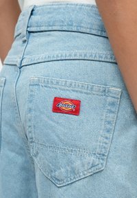 Ανοιχτό μπλε τζιν παντελόνι με πίσω τσέπη που φέρει κόκκινο και κίτρινο λογότυπο της μάρκας Dickies στη λεκάνη ενός ατόμου.