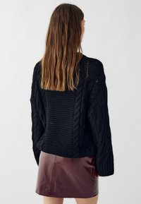 Črn pulover s kabelskim pletenjem, ohlapnega kroja in širokimi rokavi, oblečen čez sijočo burgundsko mini krilo. Teksturirana tkanina in debeli pleteninski detajli.