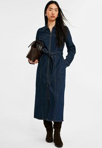 Denim-mekko, jossa on vetoketjukiinnitys edessä, pitkät hihat ja vyötärölle sidottu vö. Tumma sininen väri, jossa on kontrastoivat keltaiset ompeleet. Yhdistetty ruskean laukun ja saappaiden kanssa.