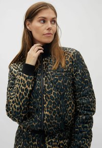 Frau mit hellbraunen Haaren, die eine gequiltete Jacke mit Leopardenmuster und schwarzen Bündchen sowie Kragen trägt, schaut nach links, Hand nahe am Gesicht.