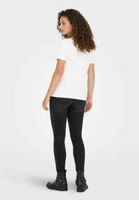 Vrouw met krullend haar, gekleed in een wit T-shirt, zwarte skinny jeans en zwarte enkellaarsjes, staand en kijkend naar links.