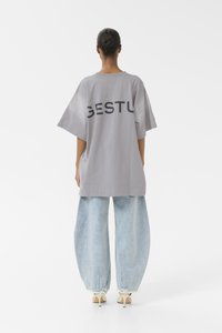 T-shirt grigia oversize con la scritta "GESTU" stampata sul retro, abbinata a jeans di denim azzurro chiaro larghi e scarpe con tacco color crema.