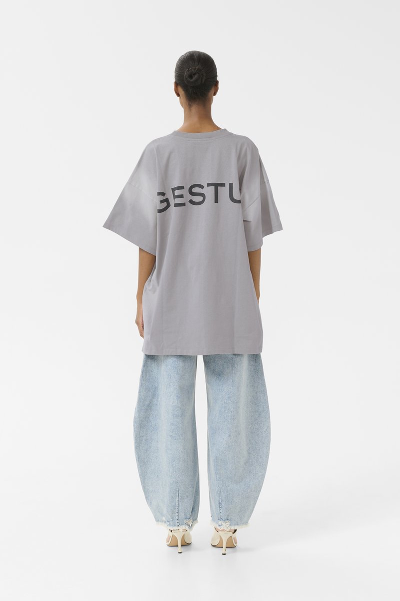T-shirt grigia oversize con la scritta "GESTU" stampata sul retro, abbinata a jeans di denim azzurro chiaro larghi e scarpe con tacco color crema.