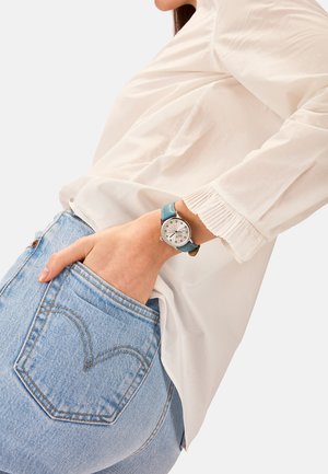 Montre avec bracelet en cuir bleu et un cadran circulaire argenté, présentant un motif texturé, des index horaires audacieux et un design minimaliste.
