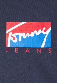 Navy-Stoff mit einem roten und blauen rechteckigen Logo, auf dem in kursiver und fetter serifenloser Schrift "Tommy JEANS" in weißer Farbe steht.