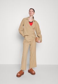 Beige wrap-paita pitkillä hihoilla ja V-pääntiellä, yhdistettyinä samansävyisiin rentoihin housuihin. Punainen ribattu toppi ja ruskeat platform-sandaalit. Kudottu laukku korostaa asua.