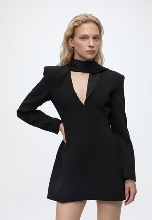 Femme aux cheveux blonds portant une robe noire à manches longues avec des épaules rembourrées et un profond décolleté en V, debout avec une main sur la hanche.
