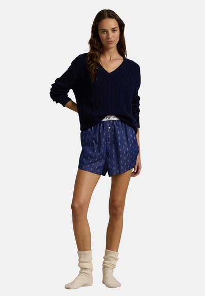 Marineblauwe kabelgebreide trui met V-hals, gecombineerd met marineblauwe shorts met een licht patroon en crèmekleurige kniekousen. Zachte texturen overal.