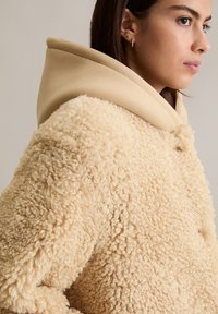 Beige pluche textuur jasje met een beige capuchon. Kenmerken zijn een ronde vorm en voorknoopjes, en het toont een zachte, donzige buitenkant.