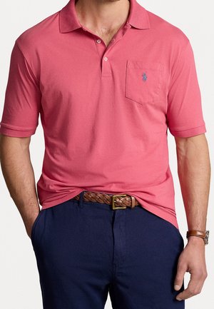 Homme portant un polo à manches courtes rose corail avec une poche sur la poitrine, une ceinture tressée marron, un pantalon bleu marine et une montre en cuir marron.