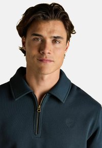 Jauns vīrietis ar brūniem matiem un riekstkoka acīm, tērpies tumšā, ar rāvējslēdzēju aizveramas apkakles džemperī uz vienkārša baltā fona.