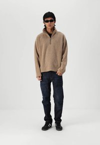 Beige rits-fleece sweatshirt met een gestructureerde afwerking, gecombineerd met donkere denim jeans en zwarte sportschoenen. Model draagt een donkere zonnebril.