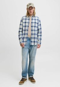 Blau-weiß kariertes Hemd, beiges Oberteil darunter, hellblaue Jeans und braune Wanderschuhe. Die Accessoires umfassen eine beigefarbene Baseballkappe.