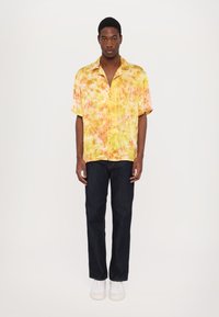 Camicia a maniche corte con bottoni, caratterizzata da un motivo tie-dye giallo e arancione, abbinata a jeans blu scuro e sneakers bianche.