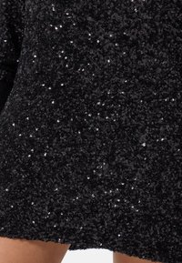 Tessuto in paillettes nere con una trama densa, caratterizzato da piccole paillettes lucide che creano una superficie scintillante. Il design include un bordo a scallop.