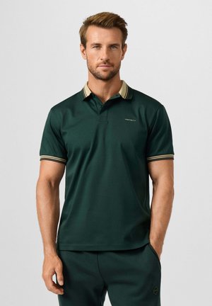 Hombre con camiseta polo de manga corta en verde oscuro con cuello y puños a rayas beige, de pie frente a un fondo claro y liso.