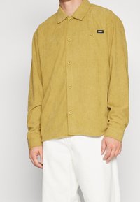Camicia button-up in corduroy senape con colletto, dotata di taschino sul lato sinistro e di una discreta toppa con logo; maniche lunghe con polsini abbottonati.