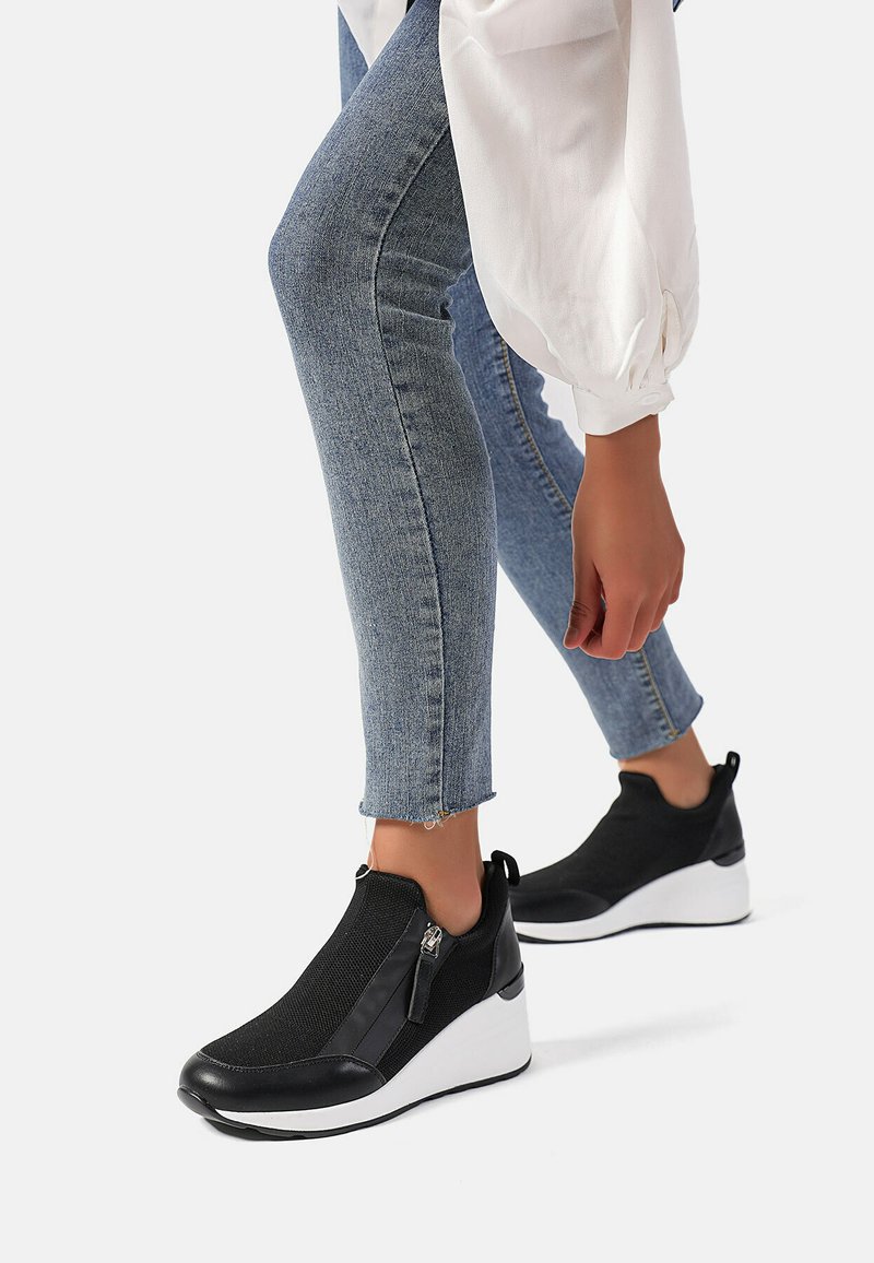 Sneaker nera con tomaia testurizzata, pannelli laterali elasticizzati e chiusura con zip. Presenta una suola a zeppa bianca e dettagli in pelle a contrasto.