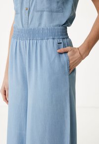 Jasnoniebieski kombinezon z chambray, z elastycznym pasem, kieszeniami bocznymi i luźnym krojem. Posiada kieszenie na guziki na piersi oraz gładką, miękką strukturę.