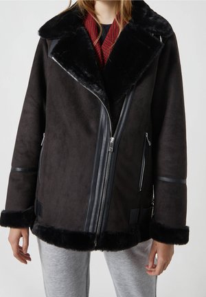 Kobieta w czarnej kurtce z kożuchem z ukośnym zamkiem błyskawicznym i czerwonym szalikiem, w połączeniu z jasnoszarymi spodniami.