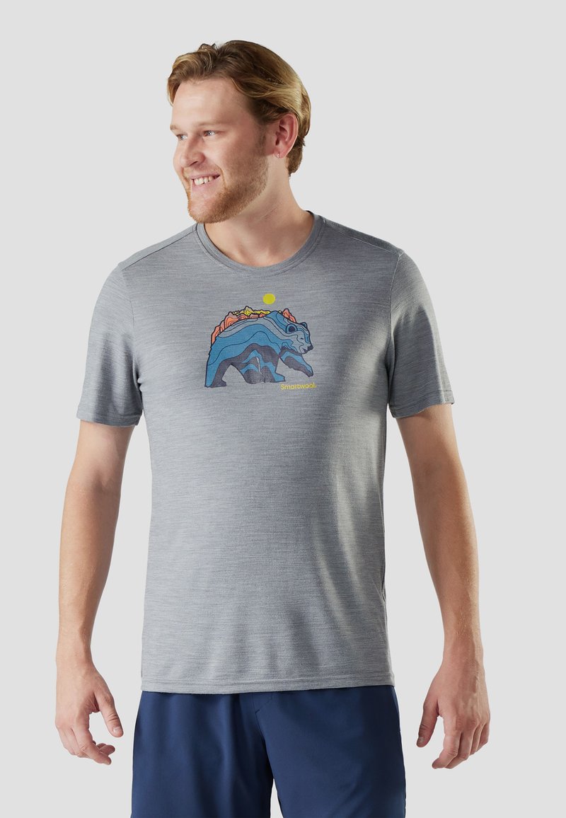 Uomo sorridente che indossa una maglietta grigia con una grafica colorata di un orso e montagne, abbinata a pantaloncini blu scuro, in piedi davanti a uno sfondo semplice.