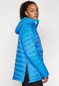 Blauwe pufferjack met een capuchon, voorzien van een gewatteerd ontwerp en een gladde afwerking. Inclusief zijritsen en een knopenaccent op de kraag.