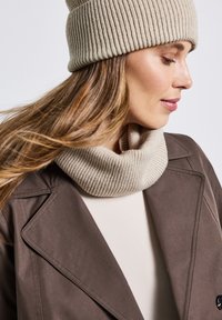 Beige gebreide beanie en bijpassende geribde col, gelaagd over een crèmekleurige top, onder een donkerbruine jas met gedetailleerde stitching.