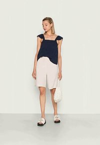 Top azul-marinho de mangas curtas com tecido texturizado, combinado com calções beiges claros. A modelo segura uma bolsa branca texturizada e usa sandálias pretas.