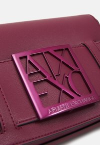 Burgundowa skórzana torebka z geometrycznym, błyszczącym różowym metalowym emblematem z wygrawerowanym napisem "ARMANI EXCHANGE". Gładka tekstura z detalami w postaci stężeń.