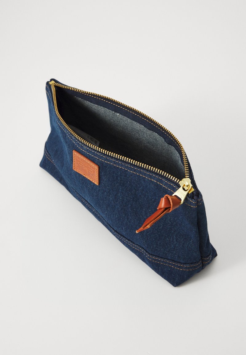 Bolsa de mezclilla con un zipper dorado, acento de cuero marrón y forro interior. Forma rectangular con detalle de costura visible.