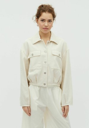 Young woman with curly hair wears a cream buttoned jacket with chest pockets and loose white pants against a plain light background.