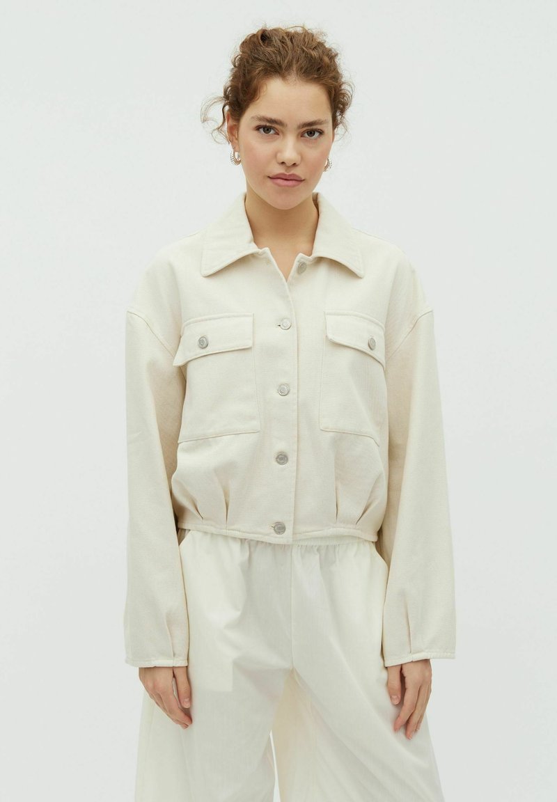 Joven con cabello rizado que lleva una chaqueta crema abotonada con bolsillos en el pecho y pantalones blancos sueltos, frente a un fondo claro y liso.