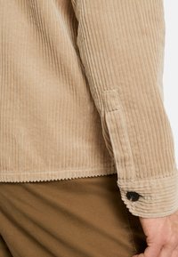 Beige Corduroy-Jacke mit vertikaler Rippung, einem knöpfbaren Manschette mit einem schwarzen Knopf und einer glatten Textur. Kombiniert mit braunen Baumwollhosen.