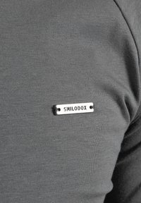Szara koszulka z materiału o gładkiej teksturze, z małą srebrną metalową metką wygrawerowaną napisem "SMILODOX" przyszytą po lewej stronie.