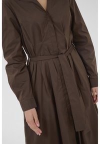 Brown long-sleeve dress featuring a V-neck, belt at the waist, and gathered fabric. The material appears smooth and lightweight.